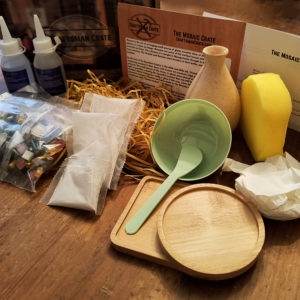 Photo of the Mosaic Crate showing adhesive, tesserae, bowl, spatula, coaster trays, wooden vase, sponge, gloves, grout mix, instructions, and splash card on a wooden table with paper fluff.