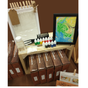 Photo of Water Marbling Crate showing Example Water Marbling Project in a frame, paints, comb tool, thickener, tube tools, tray, paper, tag, clothespins, jute string and crates sitting on a wooden box