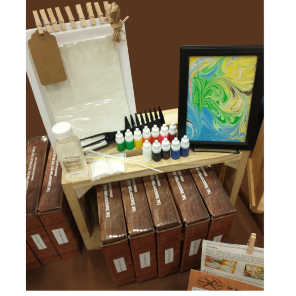 Photo of Water Marbling Crate showing Example Water Marbling Project in a frame, paints, comb tool, thickener, tube tools, tray, paper, tag, clothespins, jute string and crates sitting on a wooden box