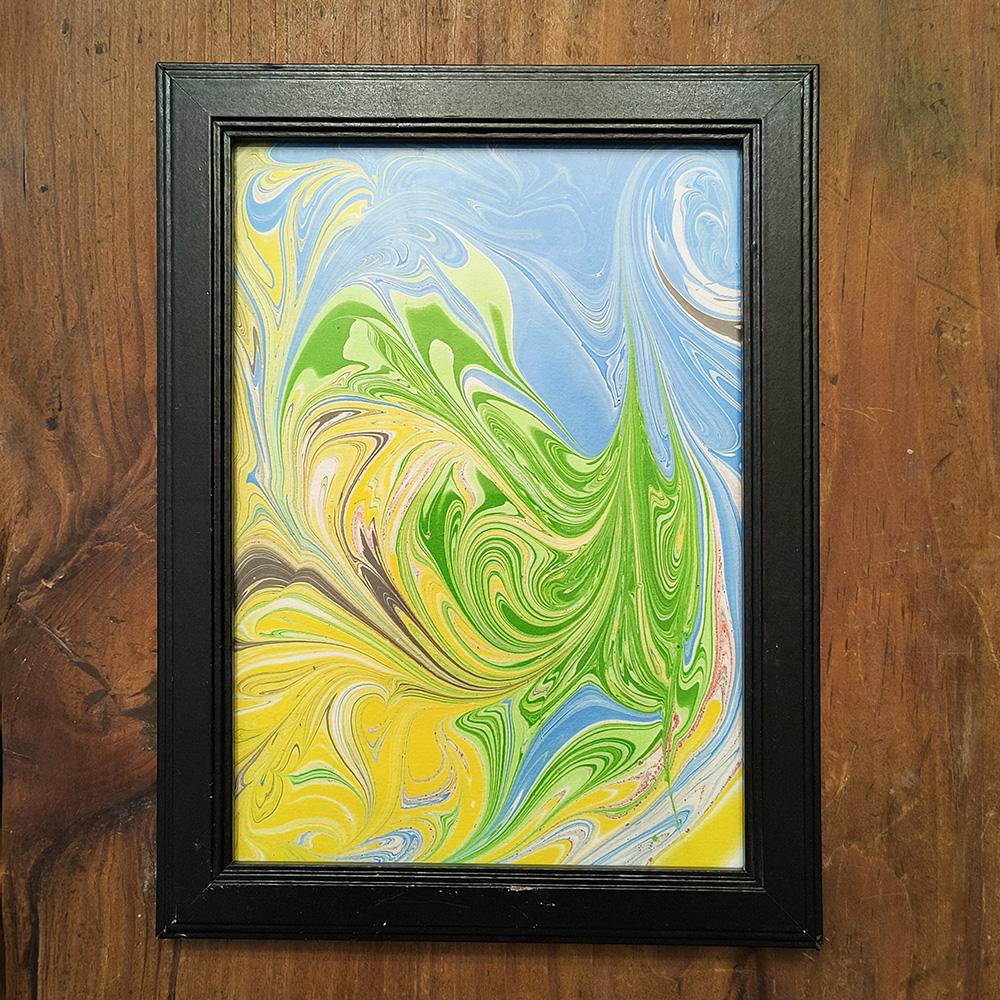 Acrylic Pouring Example An acrylic pouring painting in blues, greens, and yellows in a black frame on a wooden table.