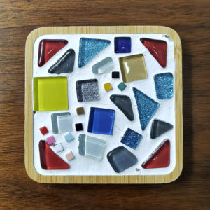 A mosaic coaster with colorful tesserae and white grout in a bamboo tray.