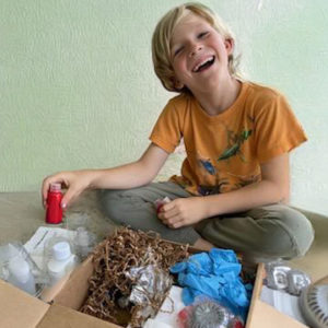 An elementary age blond boy opening the Acrylic Pouring crate. He's holding a bottle of red paint. You can see gloves, paints, canvases, and tools in the box. He has a big smile on his face. He is in front of a neutral wall.