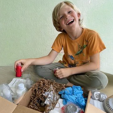 Peter Opening Acrylic Pouring An elementary age blond boy opening the Acrylic Pouring crate. He's holding a bottle of red paint. You can see gloves, paints, canvases, and tools in the box. He has a big smile on his face. He is in front of a neutral wall.