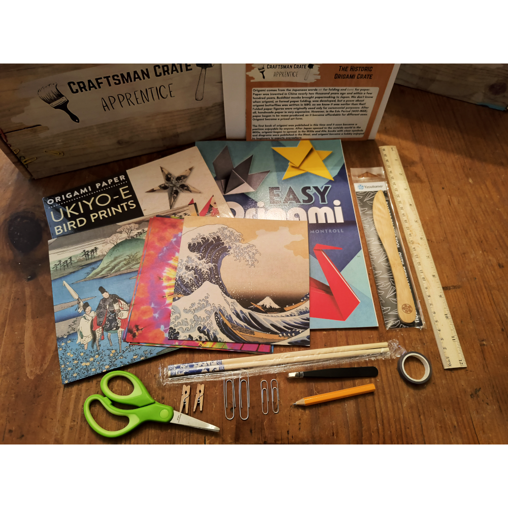 APP Origami Crate Laid Out 1000x1000 Apprentice Origami Crate laid out - Easy Origami book, Bird origami paper, three different types of loose origami paper, scissors, chopsticks, folding bone, ruler, golf pencil, tape, paper clips, mini clothespins, Apprentice box, splash card, and instructions on a wooden table.