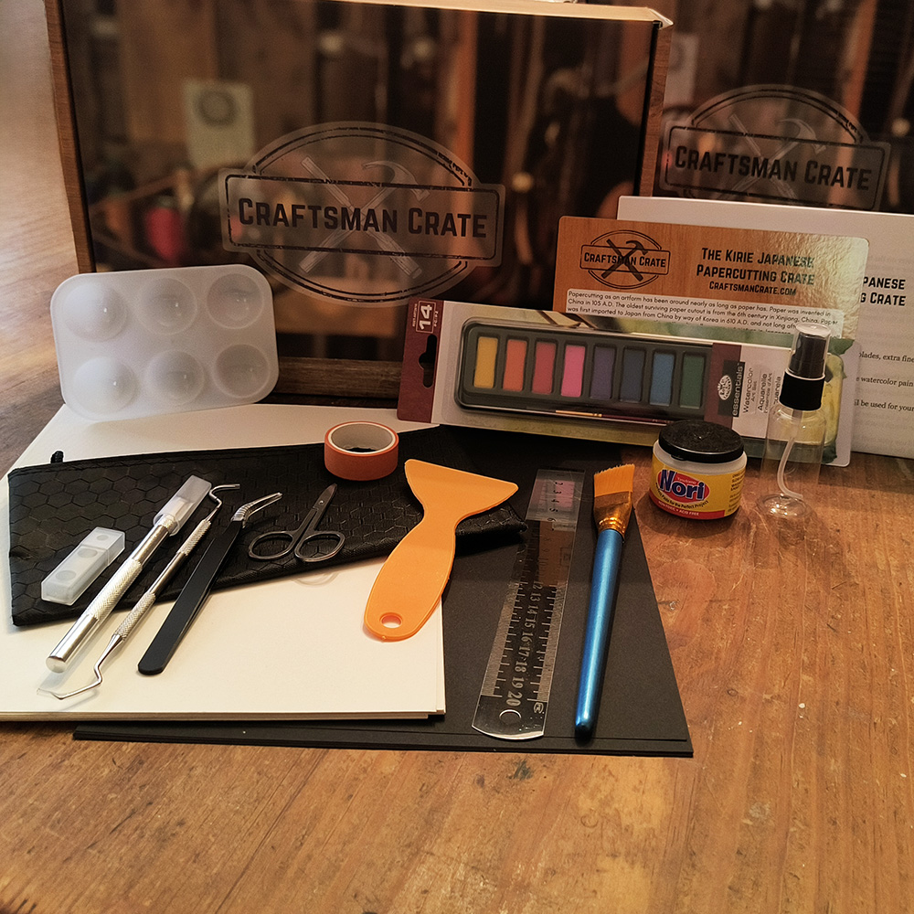 Kirie Crate Displayed 1000x1000 Craftsman Crate Kirie Japanese Papercutting Crate showing the box, watercolor palette, brush, scissors, craft knife, spatula, tweezers, metal ruler, pick, watercolor paints, splash card, instructions, black paper, watercolor paper, and tape.