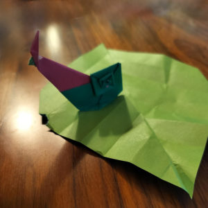 Origami snail in burgandy and dark green on an light green origami leaf on a walnut wood table.