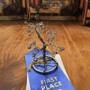 A wire sculpture tree on a wire ring sitting on top of a first place ribbon on a wooden table.